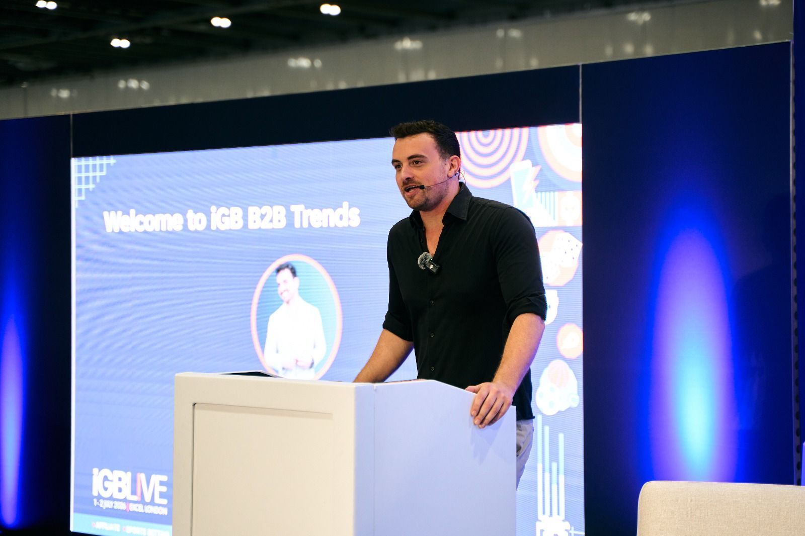 Henk Wolff speaking at a white podium at iGB Live London, the Welcome to iGB B2B Trends slide on the screen behind him.
