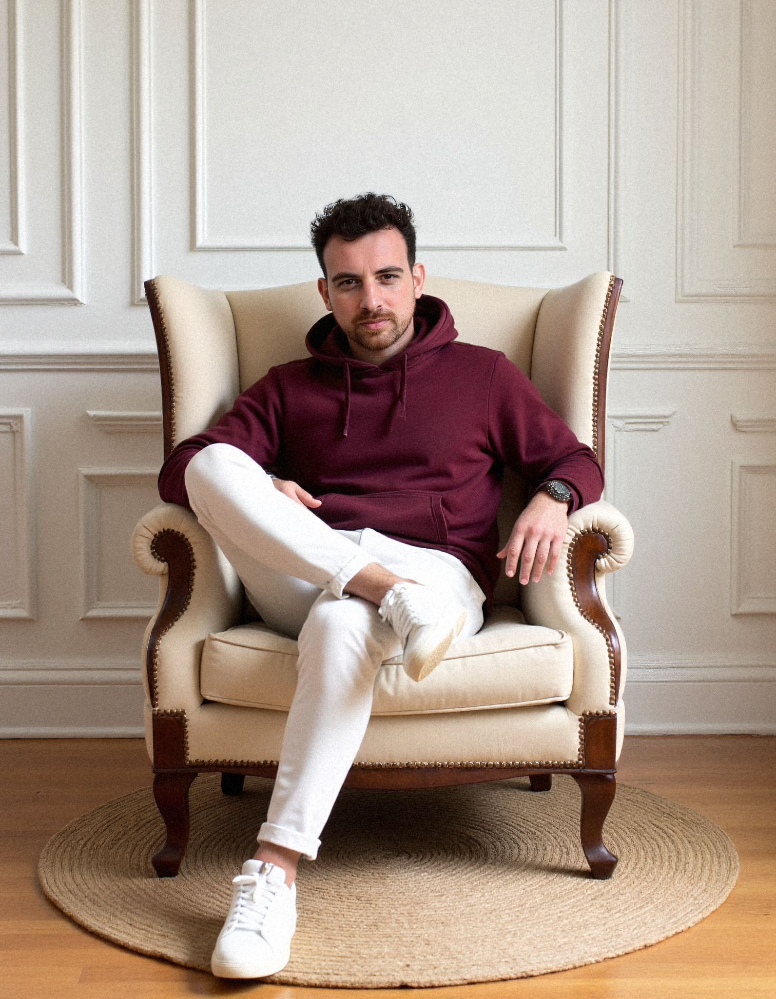 Henk Wolff seated in a cream wingback armchair with his legs crossed, against a white panelled wall.